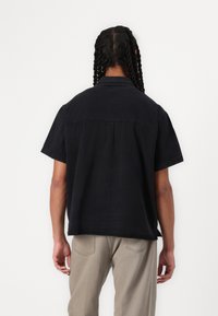 Black short-sleeved button-up shirt with a relaxed fit, featuring a back pleat for added movement and a pointed collar; fabric texture is smooth.