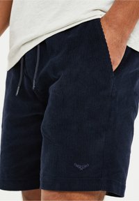 Pantalones cortos de corduroy azul marino con una superficie texturizada, cinturilla elástica, bolsillos laterales y un sutil bordado cerca del dobladillo.