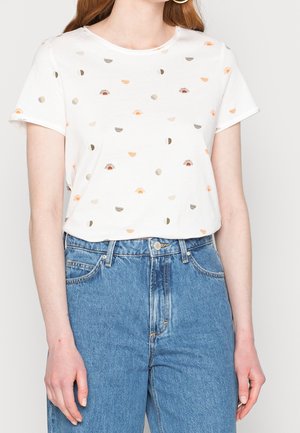 Mujer con una camiseta blanca de manga corta con pequeños patrones de sol y media luna, metida dentro de unos vaqueros de talle alto azul.