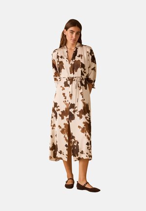 Vrouw staand in een crèmekleurige jurk tot op de knie met bruine abstracte vlekken, getailleerde taille, lange mouwen en donkerbruine platte schoenen.