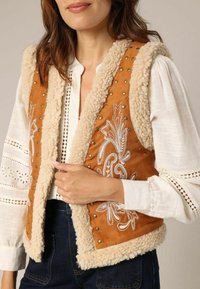 Gilet en daim marron doublé de fausse fourrure crème, avec des motifs brodés blancs et des clous métalliques. Porté par-dessus un chemisier blanc à texture.
