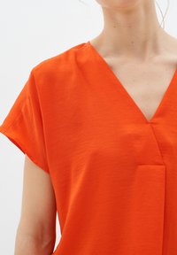 Top arancione a maniche corte con scollo a V e una finitura leggermente testurizzata. Presenta un dettaglio di cucitura frontale e una vestibilità comoda.