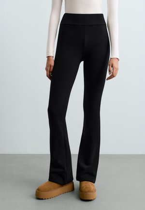 Modèle portant un pantalon noir évasé taille haute, un haut blanc et des chaussures à plateforme beige sable, sur un fond de studio uni.