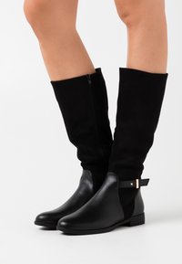 Bottes noires montant jusqu'au genou, combinant cuir et daim, avec une fermeture éclair latérale et un détail en métal doré. Semelle plate.