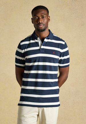 Man met een marineblauw-wit gestreept poloshirt en lichtgekleurde broek staand tegen een effen beige achtergrond.