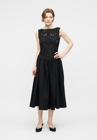 Kobieta stojąca, ubraną w czarną, bezrękawnikową sukienkę midi z haftowanymi wycięciami oraz czarne, szpiczaste buty na niskim obcasie, na białym tle.