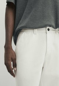 Pantaloni di cotone beige chiaro con una consistenza liscia e chiusura con bottone, abbinati a un top grigio in maglia con una vestibilità ampia.
