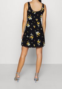 Svart ärmlös klänning med blommönster i gult och grått, utsvängd kjol och en figursydd överdel. Bärs med ljusblå sandaler med remmar.