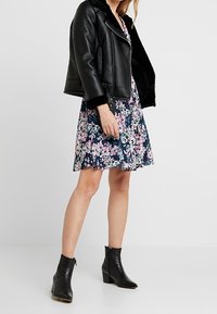 Svart lädermotorcykeljacka med fårskinnsdetaljer, lager på ett blommigt tryckklänning i marinblått, rosa och vitt, bärs med svarta anklar stövlar.