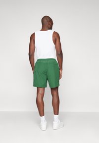 Pantalones cortos verdes de tejido ligero, con cintura elástica y un bolsillo trasero. Llevados con una camiseta sin mangas blanca y zapatillas blancas.
