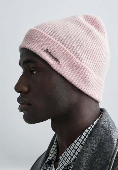 Lichtroze geribbelde muts met een omgeslagen boord, voorzien van een zilveren logo-label. De textuur lijkt zacht en gebreid, geplaatst tegen een neutrale achtergrond.