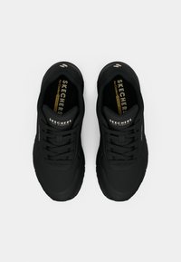 Paire de baskets noires Skechers avec lacets et semelles intérieures marquées, vues de dessus sur un fond gris clair uni.