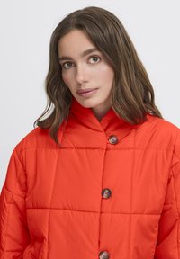 Kvinde med langt brunt hår iført en lys orange kviltet jakke med store knapper, ser roligt fremad mod en ensfarvet baggrund.