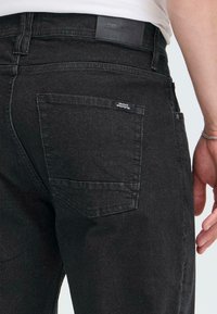 Jeans de mezclilla negra con un bolsillo trasero que tiene una pequeña etiqueta, costura visible y una textura suave. Estilo de cintura alta y corte ajustado.