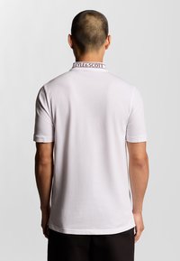 Witte polo met korte mouwen, voorzien van een ribbelkraag met "LYLE & SCOTT" branding in bordeauxrood. Gladde textuur, eenvoudig ontwerp.