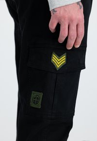 Hand mit Tattoos, die auf einer schwarzen Hosentasche mit grünen und gelben militärischen Aufnähern auf weißem Hintergrund ruht.