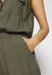 Olivenfarbener Overall mit kragenförmigem Ausschnitt, Knopfverschluss und seitlichen Taschen. Leichter Stoff mit lockerer Passform und elastischem Bund.