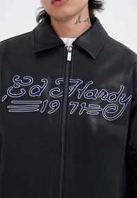 Giacca in pelle nera con zip frontale, caratterizzata da testo ricamato blu "Ed Hardy 1971" e un design di colletto classico.