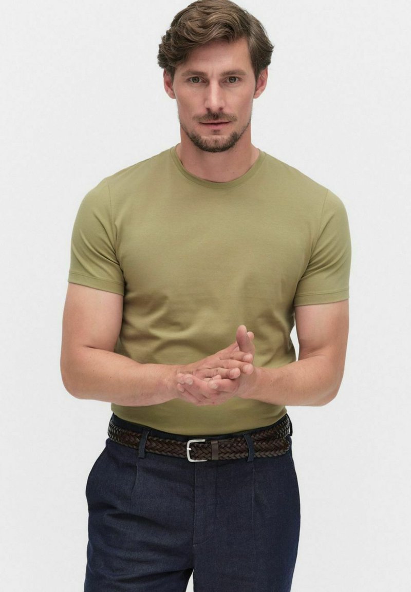 Korte mouwen, olijfgroen t-shirt gemaakt van zachte stof, gedragen met donkere marineblauwe broeken en een gevlochten bruine riem, handen gevouwen voor zich.