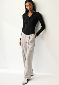 Black long-sleeve henley shirt paired with light gray pleated trousers and black-and-white athletic shoes. Plain background.