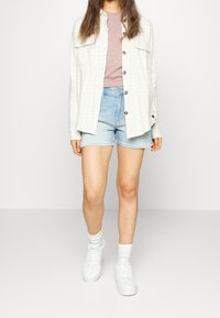 Ljus rutig skjorta med knappar över en rosa topp, kombinerad med denimshorts och vita sneakers. Outfitten har en avslappnad passform och en laid-back stil.