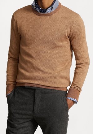 Mann trägt einen camelbraunen, strukturierten Pullover über einem gestreiften Hemd mit gepunktetem Schal, kombiniert mit dunkelgrauen Hosen, Hände in den Taschen.