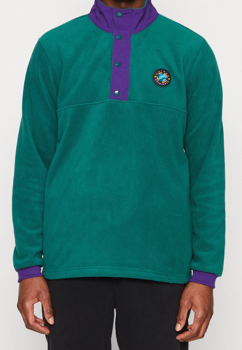 Grön fleecetröja med en lila krage, knäppt bröstslå och en cirkulär logotyp på vänstra sidan. Mjukt material och avslappnad passform.