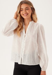 Witte, lichte blouse met bloemen uitsnijdingen, een knoopsluiting aan de voorkant en lange pofmouwen met een geruffelde kraag.