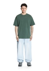 Zielony oversize'owy t-shirt z okrągłym dekoltem, noszony z jasnoniebieskimi szerokimi dżinsami. Model stoi w czarnych trampkach z białymi i szarymi akcentami.