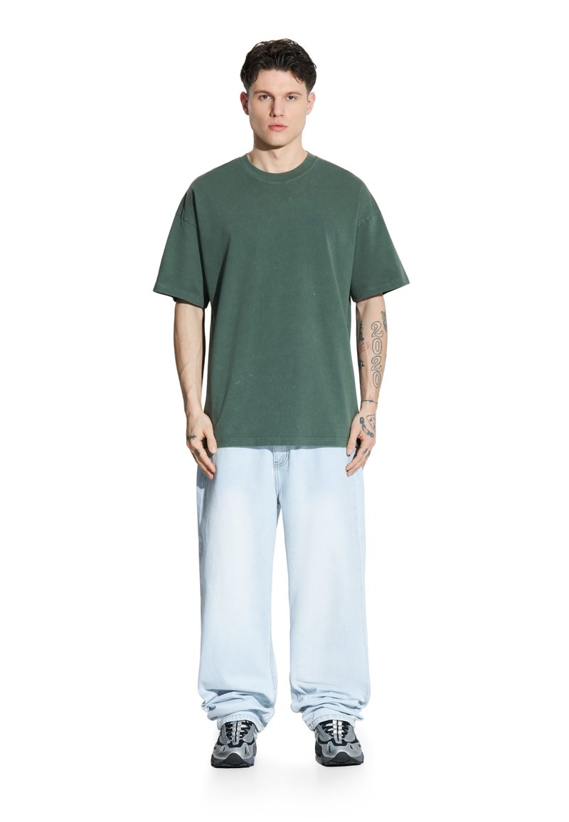 Zielony oversize'owy t-shirt z okrągłym dekoltem, noszony z jasnoniebieskimi szerokimi dżinsami. Model stoi w czarnych trampkach z białymi i szarymi akcentami.