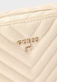 Prošívané béžové etui z umělé kůže s logem "GUESS girl" ve zlatém tónu, vybavené zipem a kontrastním prošitím.