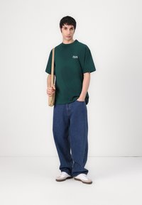 Mørkegrønn bomulls t-skjorte med korte ermer og en liten hvit logo, kombinert med vide blå denim jeans og hvite sneakers.
