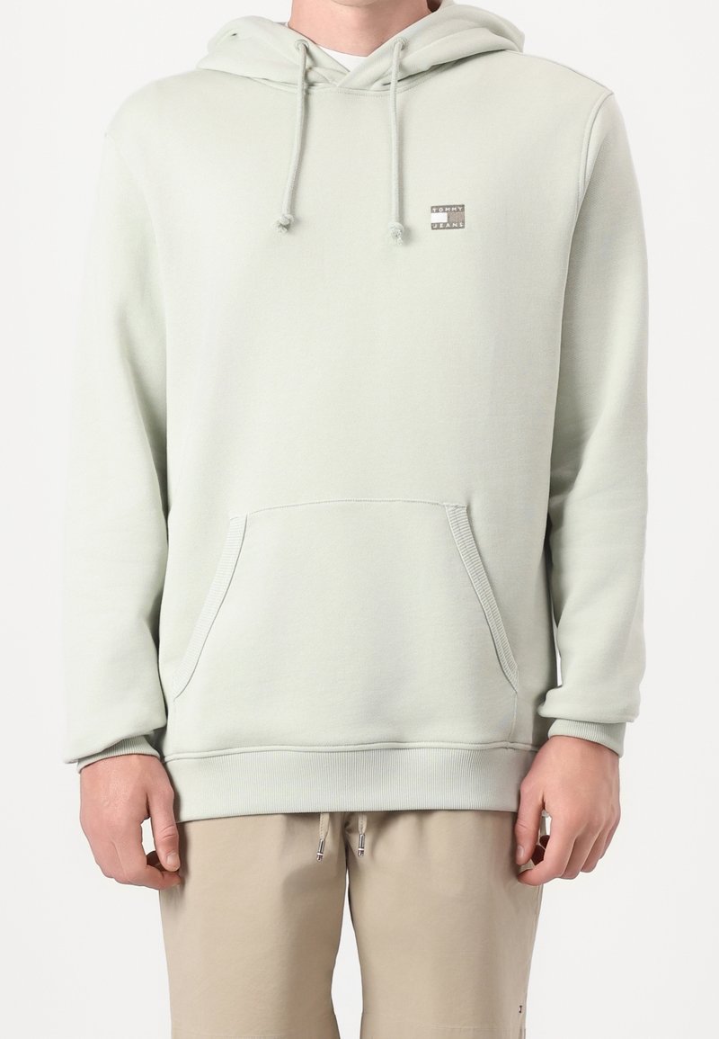 Homme portant un sweat à capuche écru avec poche avant et petit logo "Tommy Jeans" au-dessus de la poche, associé à un pantalon beige.