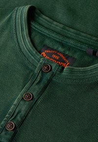 Zielona prążkowana koszulka typu Henley z trzema ciemnymi guzikami i pomarańczową metką marki wewnątrz kołnierza.