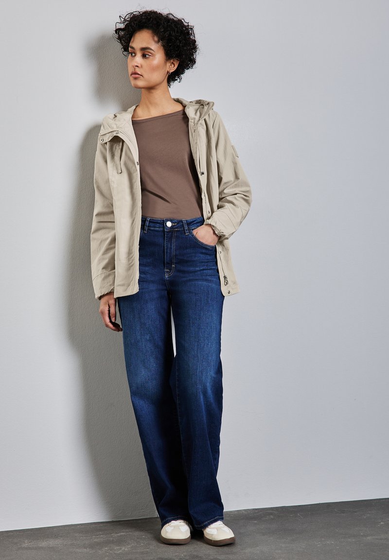 Beige zipjacka med huva, brun åtsittande topp, och vida mörkblå jeans. Vita skor med en strukturerad yta. Neutral bakgrund.