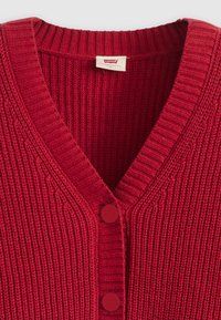 Punainen neulottu neuletakki, jossa on ribbed V-kaulus ja kaksi samansävyistä punaista nappia. Levi's-brändin etiketti kauluksessa.
