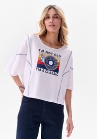 T-shirt en coton blanc avec des manches courtes et larges. Présente un imprimé graphique avec "Je ne suis pas vieux, je suis un classique", accompagné d'un design de disque vinyle.