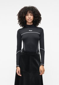 Camisola preta de manga longa com gola alta, apresentando costuras em prateado e um logo. Combinada com uma saia preta texturizada. Material liso e ajustado.