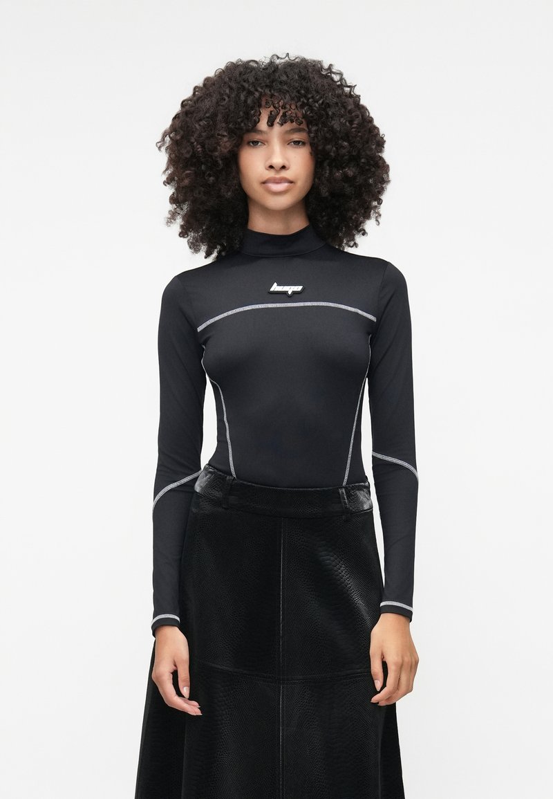 Camisola preta de manga longa com gola alta, apresentando costuras em prateado e um logo. Combinada com uma saia preta texturizada. Material liso e ajustado.