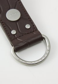 Porte-clés en cuir marron avec motif texturé, matériel en argent, anneau circulaire et détail de branding en relief. Comprend des rivets pour un détail supplémentaire.