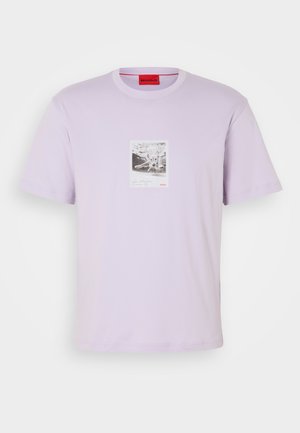 Gaiši lavandas t-krekls no kokvilnas ar centrālo grafiku, kurā attēlots astoņkājis melnbaltā krāsā, ar sarkaniem logo akcentiem.