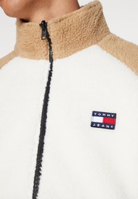 Valkoinen fleece-takki vaaleilla korostuksilla ja korkealla kauluksella. Varustettu mustalla vetoketjulla ja pienellä "TOMMY JEANS" -logolla rinnassa.