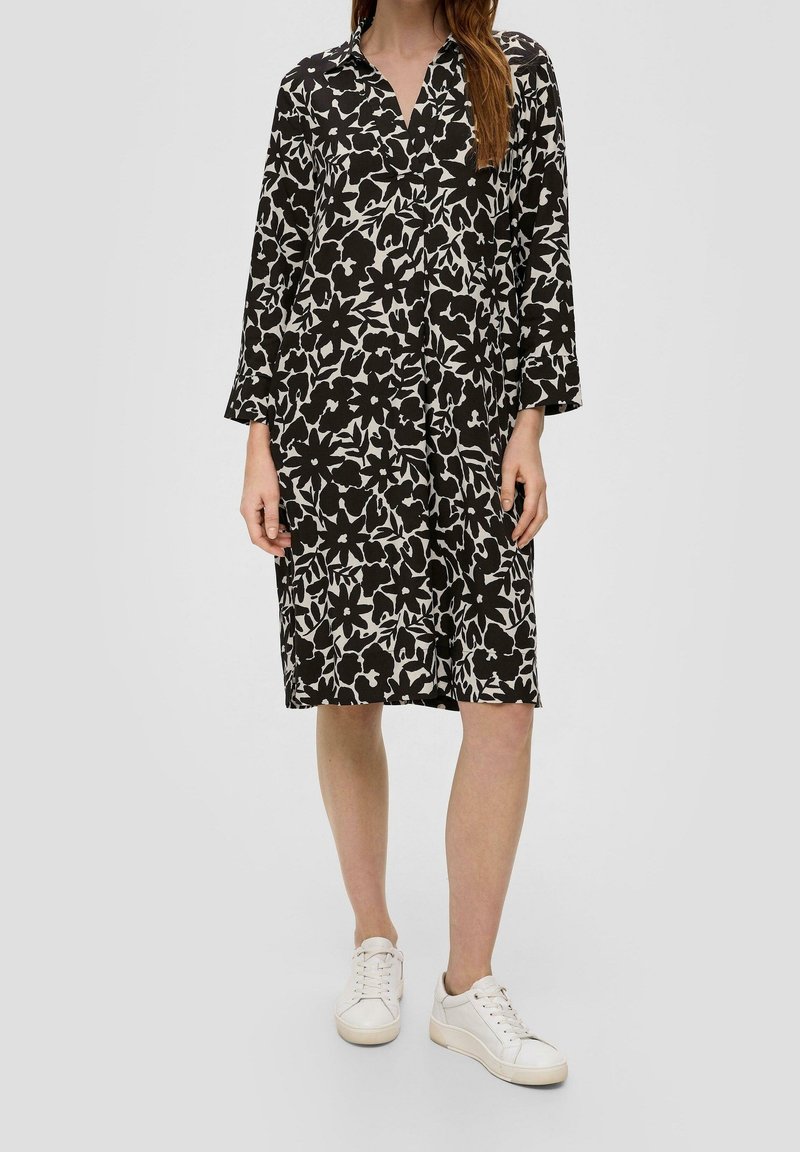 Femme portant une robe mi-longue noire et blanche à motifs floraux, manches longues, et des baskets blanches, debout devant un fond uni.