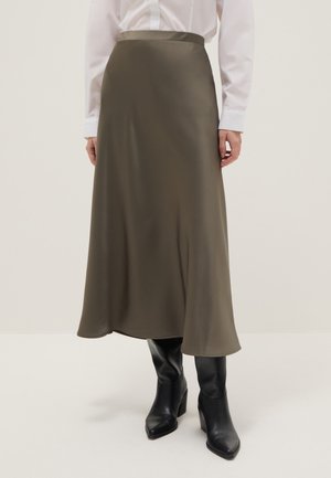 Frau trägt einen olivgrünen Midirock kombiniert mit schwarzen Stiefeln mit Absatz und einem weißen Langarmshirt, steht vor einem einfarbigen Hintergrund.