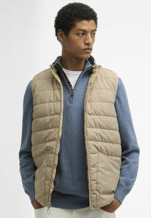 Jonge man die een beige gewatteerde bodywarmer draagt over een blauwe trui met rits, staat met zijn handen in zijn zakken en kijkt opzij tegen een witte achtergrond.