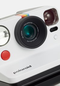 Appareil photo Polaroid blanc avec un objectif noir. Présente un bouton rouge, une étiquette de mise au point automatique et une bande colorée. Inclut un viseur et un flash.