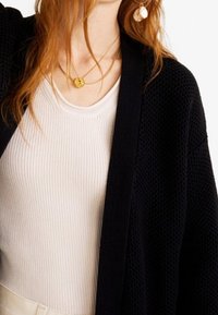 Un cardigan en tricot noir avec une coupe décontractée, superposé à un haut côtelé beige clair. Un collier en or est visible, avec un matériau texturé et un design à l'avant ouvert.