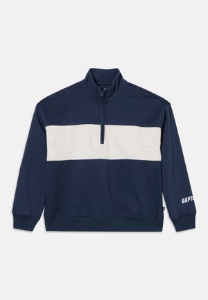 Pull bleu marine avec une bande horizontale blanche sur la poitrine, une fermeture éclair quart de longueur, et le logo GAP sur le poignet de la manche gauche.