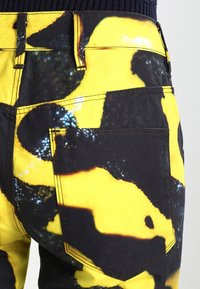 Gula och svarta mönstrade denimbyxor med en bakficka, synliga sömmar och en slät struktur. Designen påminner om abstrakta former.