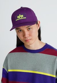 Lila sapka lapos karimával, fekete folttal, rajta "ALPHA INDUSTRIES" felirattal zöld színben. A modellen csíkos, több színben játszó pulóver van.